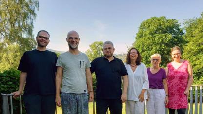Auf einer grau gepflasterten Terasse stehen sechs l&auml;chelnde Personen vor einer gr&uuml;nen H&uuml;gellandschaft mit B&auml;umen und Wiese (dem Taunus). Sie l&auml;cheln in die Kamera. Die Personen sind sommerlich gekleidet.