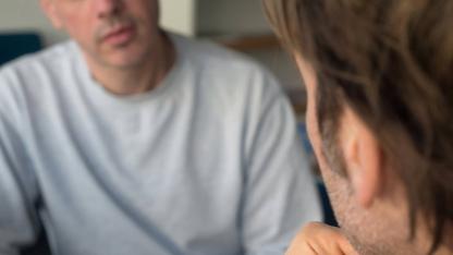 Zwei männlich gelesene Personen sitzen sich in einem ernsten Gespräch gegenüber.