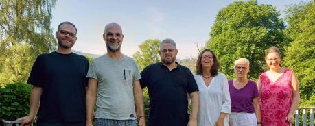 Auf einer grau gepflasterten Terasse stehen sechs l&auml;chelnde Personen vor einer gr&uuml;nen H&uuml;gellandschaft mit B&auml;umen und Wiese (dem Taunus). Sie l&auml;cheln in die Kamera. Die Personen sind sommerlich gekleidet.