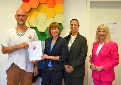 Übergabe des Förderbescheids. Vier lächelnde bzw. freundlich blickende Personen in einem Innenraum vor einer künstlerischen Plastik in Farben der Regenbogenfahne, die an einer Wand installiert ist. Die Person in der Mitte (Ministerin Hofmann) übergibt der Person ganz links (Aidshilfe-Vorstand Martí Klatt) ein Dokument, auf dessen Titel das Wappen des Landes Hessen abgebildet ist.