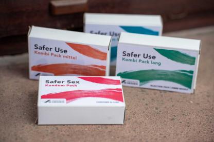 Zu sehen sind vier sogenannte "Safer-Use-Packs" in unterschiedlichen farblichen Gestaltungen: Kleine P&auml;ckchen, die Utensilien f&uuml;r den Infektionsschutz enthalten.