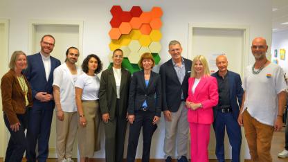 Zehn lächelnde, freundliche Personen stehen als Gruppe in einem Innenraum vor dem an einer Wand installierten plastischen Kunstobjekt in Farben der sogenannten Regenbogenfahne. In der Mitte die Hessische Ministerin für Soziales, Heike Hofmann, und die Hessische Antidiskriminierungsbeauftragte Berivan Şekerci.